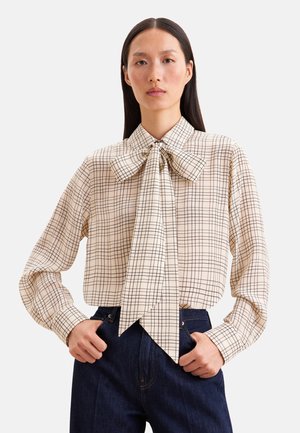 Kvinde iført en beige bluse med lange ærmer med sort ternet mønster og stor sløjfe foran, kombineret med mørkeblå højttaljede jeans.