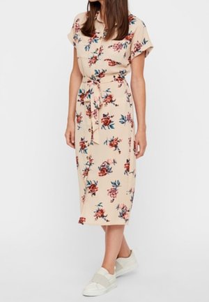 Nainen beige midimekko päällä, jossa punaisia ja sinisiä kukkakuvioita, lyhyet hihat, vyötärökaitale ja valkoiset slip-on-tennarit.