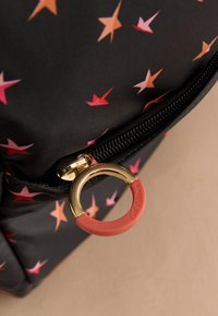 Juoda audinio medžiaga su raudonų ir rožinių žvaigždžių raštu, arti auksinės užtrauktuko traukimo kilpos, iš dalies padengtos oranžiniu guminio dangteliu, ant smėlio spalvos fono.