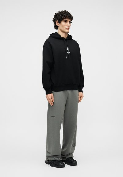 Νεαρός άνδρας με σγουρά μαλλιά, φορώντας μαύρο φούτερ με κουκούλα και χαλαρό γκρι παντελόνι φόρμας, στέκεται μπροστά σε ένα απλό φωτεινό φόντο.