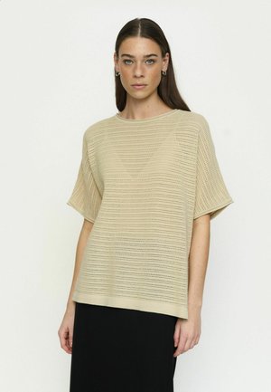 Młoda kobieta z długimi brązowymi włosami w beżowej, krótkiego rękawa, perforowanej dzianinowej bluzce i czarnej spódnicy, stoi na tle jasnego, jednolitego tła.