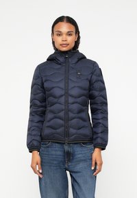 Tumši zils pūkainais apmetnis ar quilted dizainu, kapuci un priekšējo rāvējslēdzēju. Aprīkots ar mazu logotipu uz krūšu un ribotām piedurknēm.