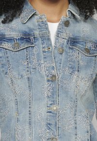 Vaaleansininen farkkutakki, jossa on koristeellista valkoista kukkakirjailua. Varustettu metallinapeilla ja klassisella kauluksella. Kaksi povitaskua näkyvissä.