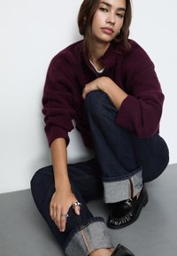 Bordo pulover iz mešanice volne s velikimi gumbi, v kombinaciji s temnimi jeans hlačami s zavihanimi sivo oblačili in črnimi loafer čevlji.