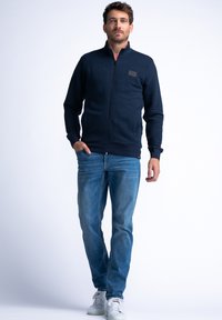 Navy zip-up jacket with ribbed texture and a small logo patch, paired with light blue jeans and white sneakers. Simple, casual design.