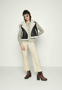 Colete de pelúcia preto e bege por cima de uma blusa verde-claro, combinado com calças largas beigas e botins castanhos. Acessório com óculos de sol brancos.