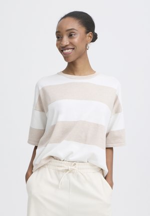 Lächelnde Frau mit Dutt, trägt einen kurzärmligen Pullover mit breiten beigen und weißen Streifen und cremefarbene Hosen mit Tunnelzug, die Hände in den Taschen.