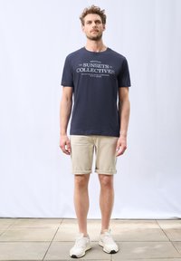 Granatowa bawełniana koszulka z nadrukiem "SUNSETS COLLECTIVES" w białym kolorze. Beżowe szorty z podwiniętymi nogawkami, jasne trampki z teksturowanym wzorem, stojące na kamiennej podłodze.