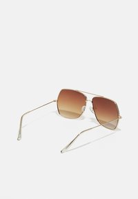 Gafas de sol de metal dorado con un marco cuadrado, lentes marrón degradado y puntas transparentes en las varillas. Presentan almohadillas nasales ajustables.