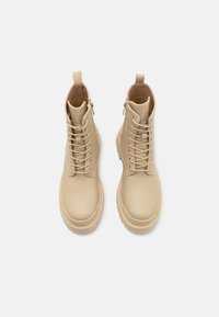 Botas de tobillo beige con cordones, con un empeine de cuero suave, puntera redonda y suela de goma gruesa; presenta una cremallera lateral y una tira para facilitar su uso.