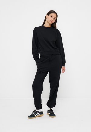 Frau posiert in lässigem komplett schwarzem Outfit mit Sweatshirt, Jogginghose, weißen Socken und schwarzen Adidas-Sneakers mit weißen Streifen.