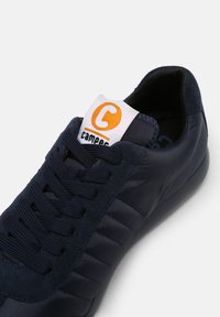 Sötétkék sportcipő sima textil felsőrésszel, velúr díszítéssel és fűzős kialakítással. Fehér címke található rajta, kör alakú narancssárga logóval.