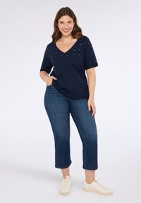 Tummansininen V-kaula-aukkoinen t-paita, jossa on värikkäitä koristeita kauluksessa, yhdistettynä tummansinisiin 7/8-farkkuihin ja vaaleisiin lenkkareihin, seisontaposeeraus.