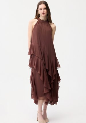 Junge Frau in einem ärmellosen, braunen, mit Volants versehenen Midikleid mit Neckholder und beigen Knöchelriemen-Pumps vor weißem Hintergrund.