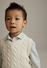 Gilet en maille torsadée blanche sur une chemise à col rayée bleu clair et blanche. Cheveux courts et foncés, avec un fond neutre.
