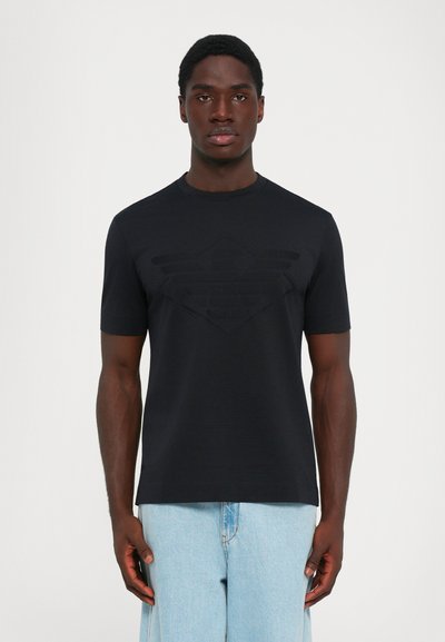 Uomo che indossa una t-shirt nera aderente con un logo in rilievo sottile e jeans azzurri chiari, in piedi contro uno sfondo bianco semplice.