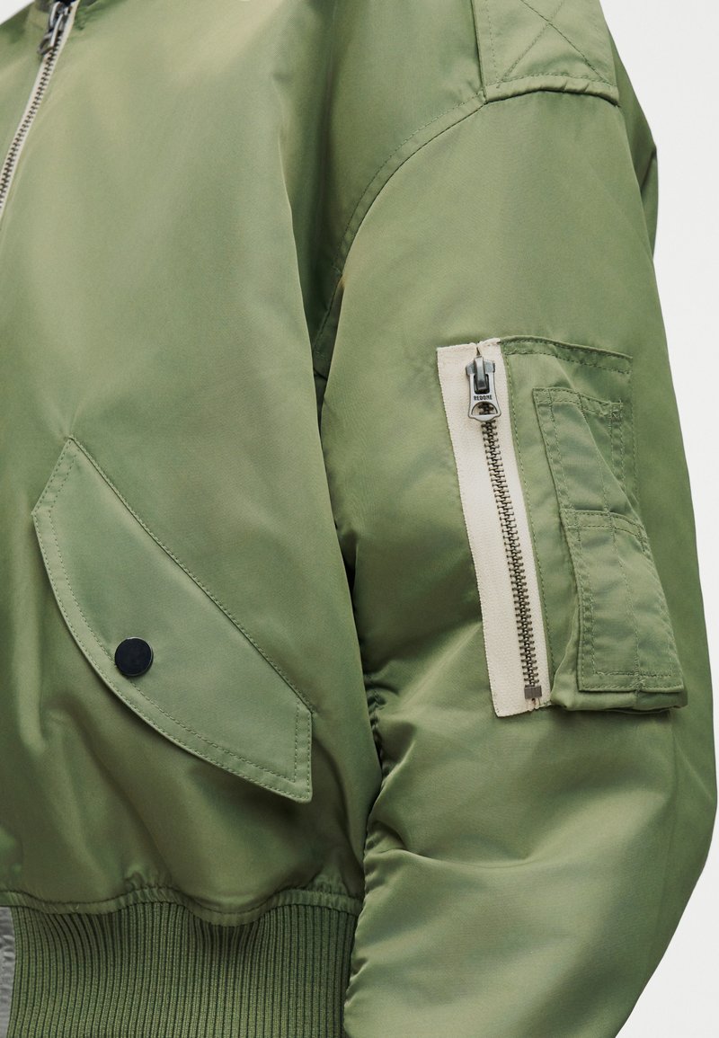 Manche de veste bomber vert olive avec poche zippée garnie de biais blanc et poche à rabat, portée sur un poignet côtelé.