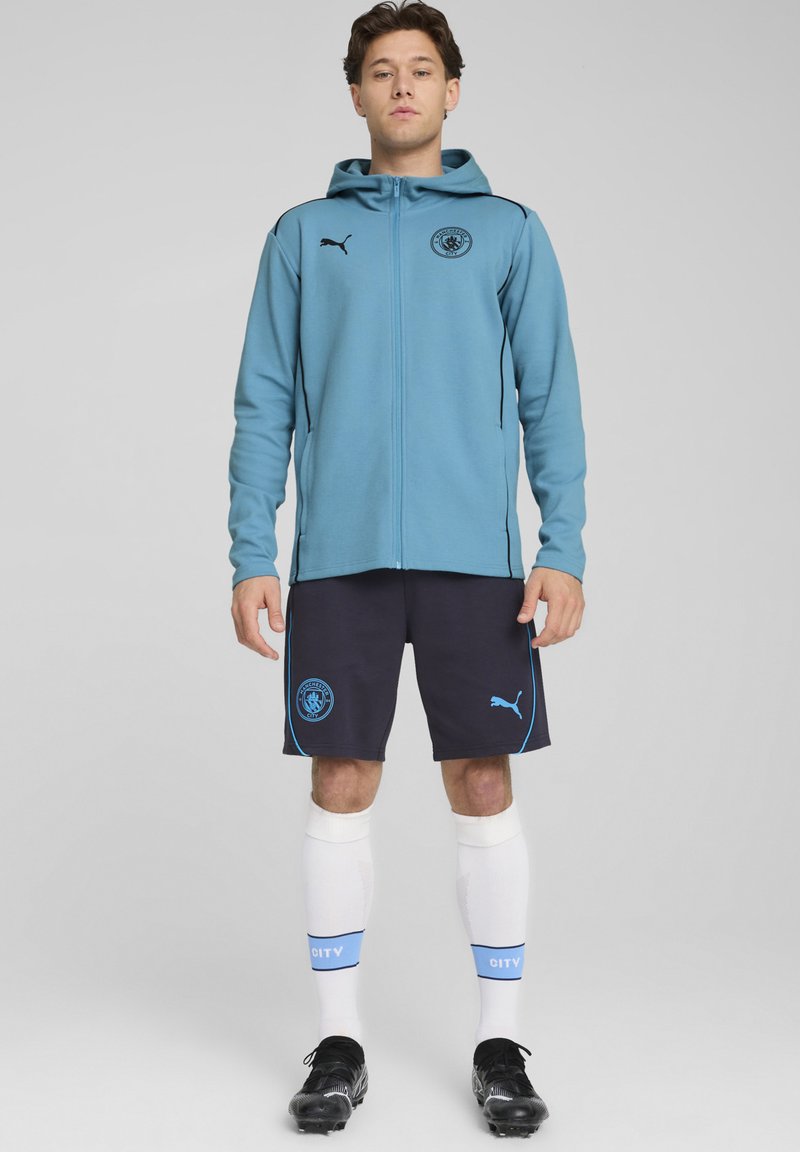 Lyseblå lynlås hættejakke med sorte accenter og logo, parret med marineblå shorts med blå syninger og logo. Hvide sokker med blå bånd.