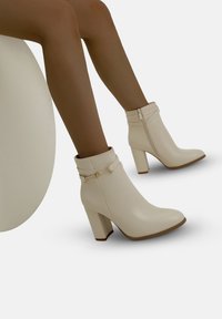 Ben klädda i beige högklackade ankelstövlar med sidodragkedja och dekorativ rem, mot en enfärgad vit bakgrund.