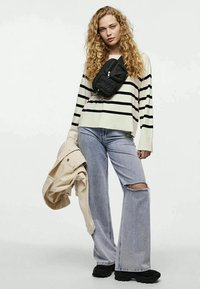Mulher com cabelo encaracolado, vestindo um suéter às riscas, jeans largos rasgados, sapatilhas pretas, bolsa transversal, segurando uma jaqueta bege.