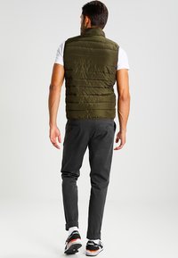 Olivgrüne Pufferweste mit horizontaler Steppstruktur, hohem Kragen, getragen über einem weißen T-Shirt, kombiniert mit dunklen Hosen und sportlichen Schuhen.