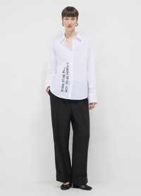 Chemise blanche à boutons avec du texte noir "NÉ POUR ÊTRE RÉEL, PAS POUR ÊTRE PARFAIT" sur le côté ; associée à un pantalon noir ample et des chaussures plates.