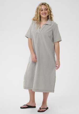 Kobieta z kręconymi włosami, ubrana w luźną, krótką, paskowaną sukienkę midi i czarne japonki, stojąca na jednolitym tle.