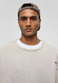 Beige oversized sweatshirt met een versleten halslijn, gedragen over een wit T-shirt, gecombineerd met een lichtbruine pet. Inclusief een oorbel.