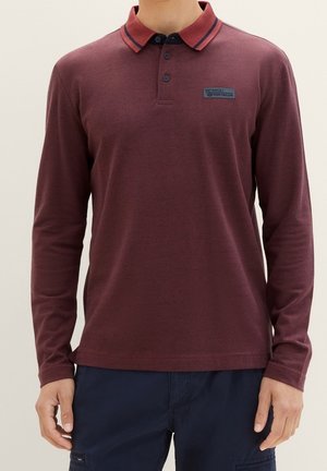 Mies, jolla on päällään marsalanpunainen pitkähihainen poolopaita, jossa on raidallinen kaulus, ja tummansiniset housut, seisomassa yksiväristä taustaa vasten.