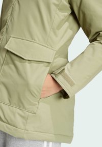 Světlá zelená izolační bunda s velkými předními kapsami, zipem a akcenty na manžetách z suchého zipu. Hladký materiál se šitými švy.