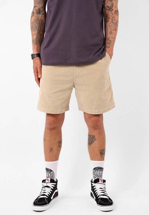 Shorts en velours côtelé beige à coupe droite, portés avec des chaussures montantes noires et des chaussettes blanches comportant un logo graphique.