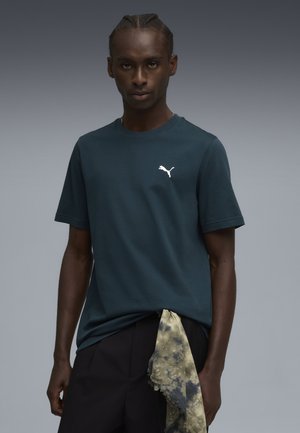 Dunkelblau-grünes T-Shirt mit kurzen Ärmeln aus Baumwolle, mit einem weißen Puma-Logo auf der Brust und einem geknoteten, gemusterten Schal an der Taille.