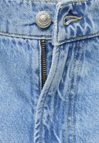 Tessuto di denim azzurro chiaro con cuciture visibili, dotato di un bottone argento e una chiusura a zip. La texture appare morbida con un aspetto leggermente vissuto.