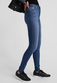 Vaqueros de mezclilla azul ajustados con un corte de talle medio, costuras detalladas y bolsillos. Combinados con zapatillas negras y una chaqueta de cuero negra.