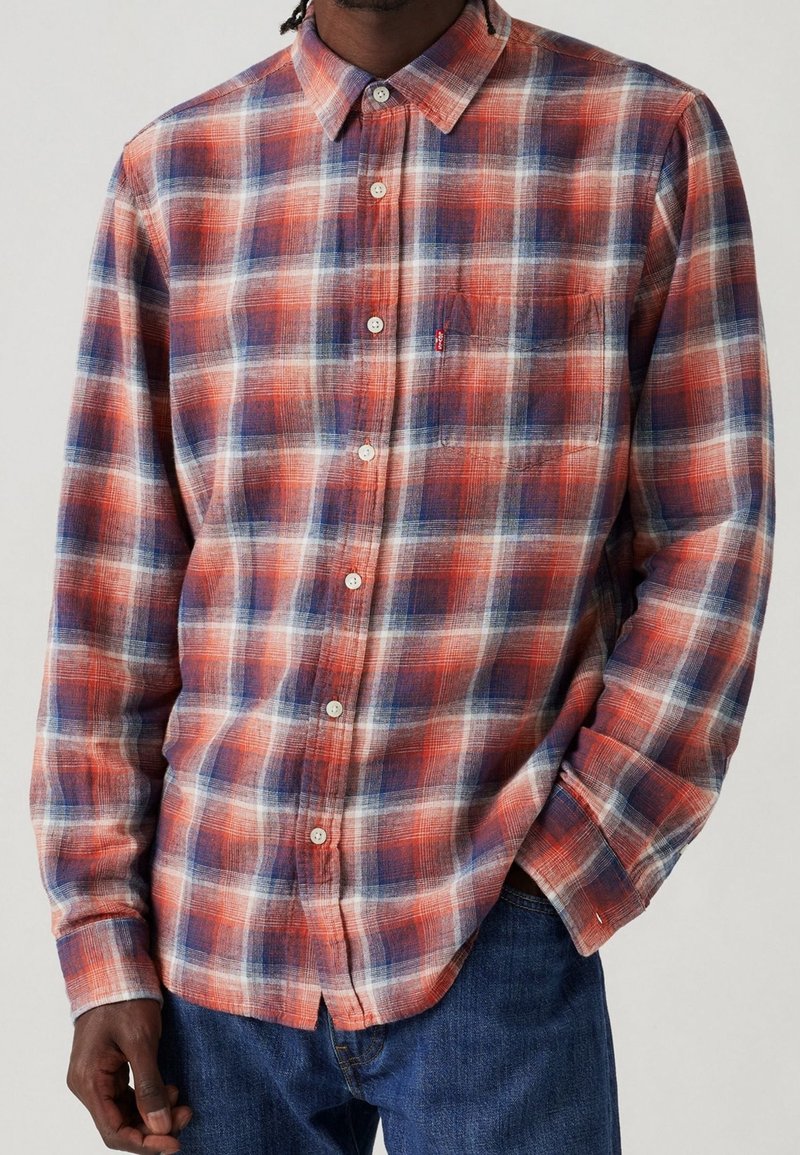 Homme portant une chemise à carreaux rouge, blanche et bleue à manches longues avec poche poitrine, associée à un jean bleu, debout devant un fond uni.