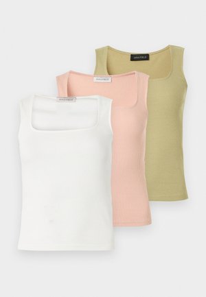 Kvinde iført khaki firkantet nakketop, brede blå jeans med forlommer, hvide hæle og mørke solbriller, stående op ad en hvid væg.