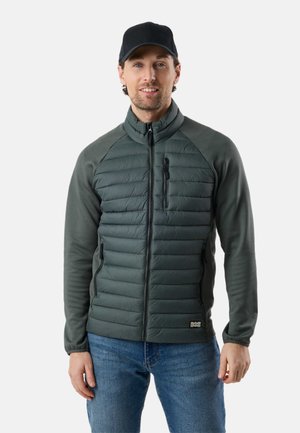 Man wearing black cap, green quilted jacket with zippered pockets, and blue jeans, standing against a plain light background.