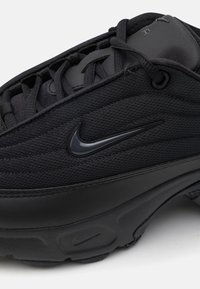 Des baskets Nike noires présentent une tige en maille texturée, des accents en plastique, des lacets ronds et une semelle en caoutchouc avec un logo Nike sur le côté.