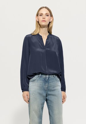 Navyblaue Satinbluse mit V-Ausschnitt, langen Ärmeln und gerafften Manschetten; kombiniert mit hellblauen, ausgewaschenen Jeans.