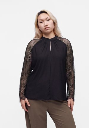 Femme aux cheveux blond clair portant un haut noir à manches longues en dentelle transparente et un pantalon marron, debout devant un fond blanc uni.
