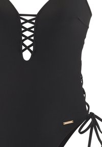 Μαύρο ολόσωμο μαγιό με βαθύ V-λαιμόκοψη και σχέδιο σταυρωτών λεπτομερειών, πλαϊνές γραβάτες και μια μικρή χρυσή ετικέτα. Λείος, ελαστικός υλικός.