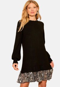 Schwarzes gestricktes Kleid mit langen Puffärmeln, gerippten Bündchen und einem schimmernden, mit Pailletten besetzten Rüschensaum. Die Textur kontrastiert zwischen Strick und Pailletten.