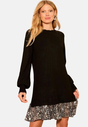 Rochie neagră tricotată cu mâneci lungi bufante, manșete ondulate și un tiv cu volan din paiete strălucitoare. Textura contrastează între tricot și paiete.