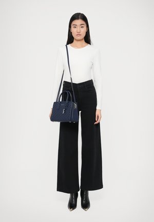 Τσαντάκι από δέρμα σε ναυτικό μπλε με διακοσμητικές διάτρητες λεπτομέρειες, κοντά χερούλια και αποσπώμενο ιμάντα ώμου, σε συνδυασμό με εφαρμοστή λευκή μπλούζα και μαύρο παντελόνι φαρδιάς γραμμής.