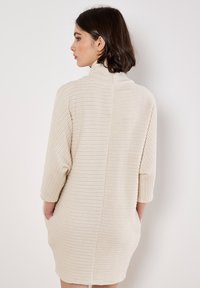 Vestito maglione color beige chiaro con un design testurizzato, maniche 3/4 e tasche laterali. Presenta un colletto alto e una cucitura dritta sul retro.
