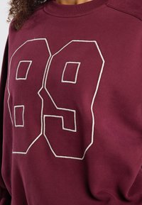 Bordeaux sweatshirt met wit, omrande cijfers "89" prominent op de voorkant. De stof is glad, met ribbelboord aan de kraag en manchetten.