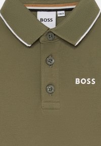 Olivgrön polo med texturerad tyg, vit krage, tre grå knappar och "BOSS"-logotyp i vitt på framsidan.