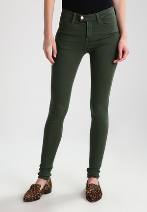 Smaltsiddende mørkegrønne bukser båret med loafers i leopardprint og en sort top, vist fra taljen til fødderne mod en ensfarvet baggrund.