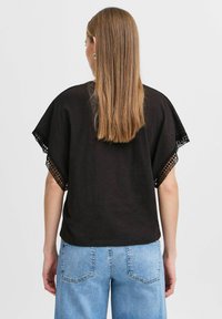 Zwarte top met korte mouwen, voorzien van kant aan de randen. De stof lijkt lichtgewicht en heeft een ontspannen pasvorm. Gecombineerd met blauwe denim jeans.