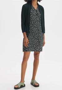 Femme portant une robe à pois noir et blanc, un cardigan ouvert vert foncé et des sandals vertes à semelles noires épaisses, se tenant sur un fond blanc.